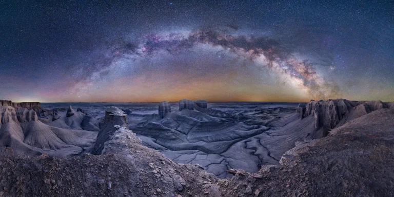 A panoramic night‑sky photograph showing the Milky Way arching above the barren, lunar-like landscape of Moonscape Overlook in Utah, captured from a vantage point.
