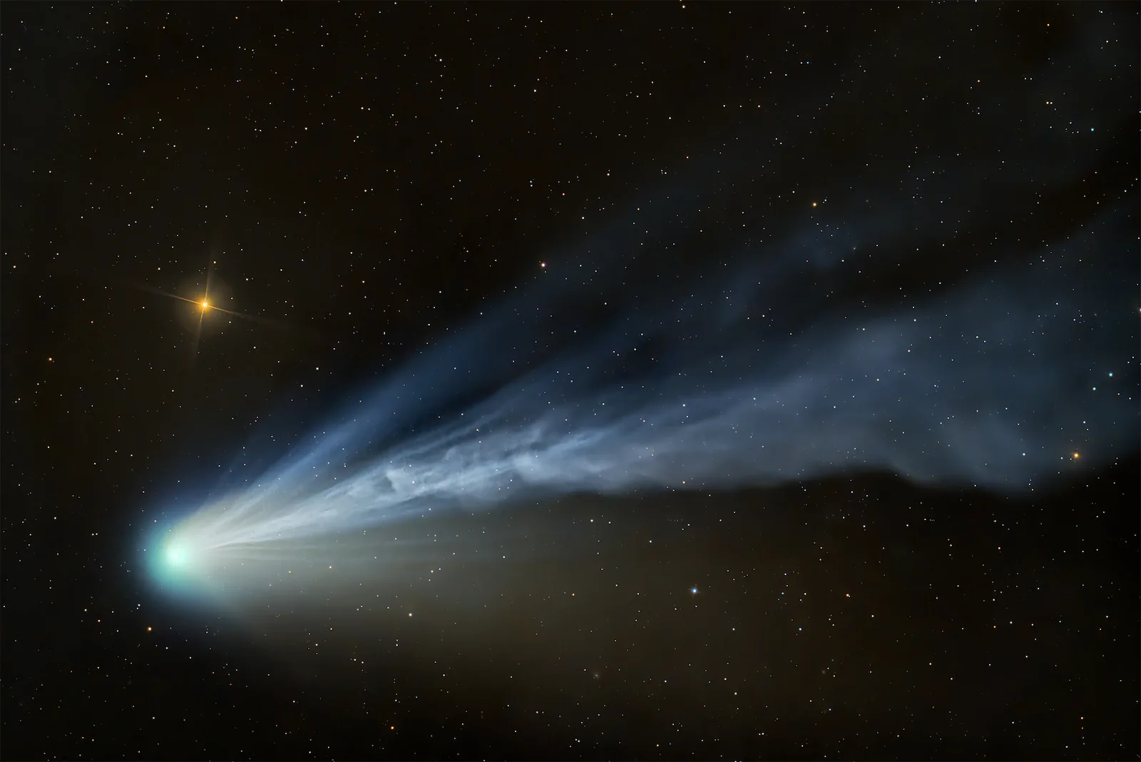 An image showing Comet 12P/Pons‑Brooks with a flowing, wind‑shaped tail, its blue tones contrasting with the warm glow of a nearby star.
