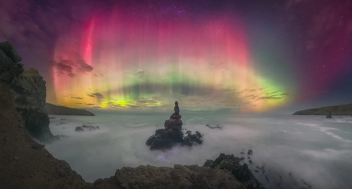 An image showing a rocky shoreline at night with a circular aurora in vivid pinks yellows and greens lighting up the sky